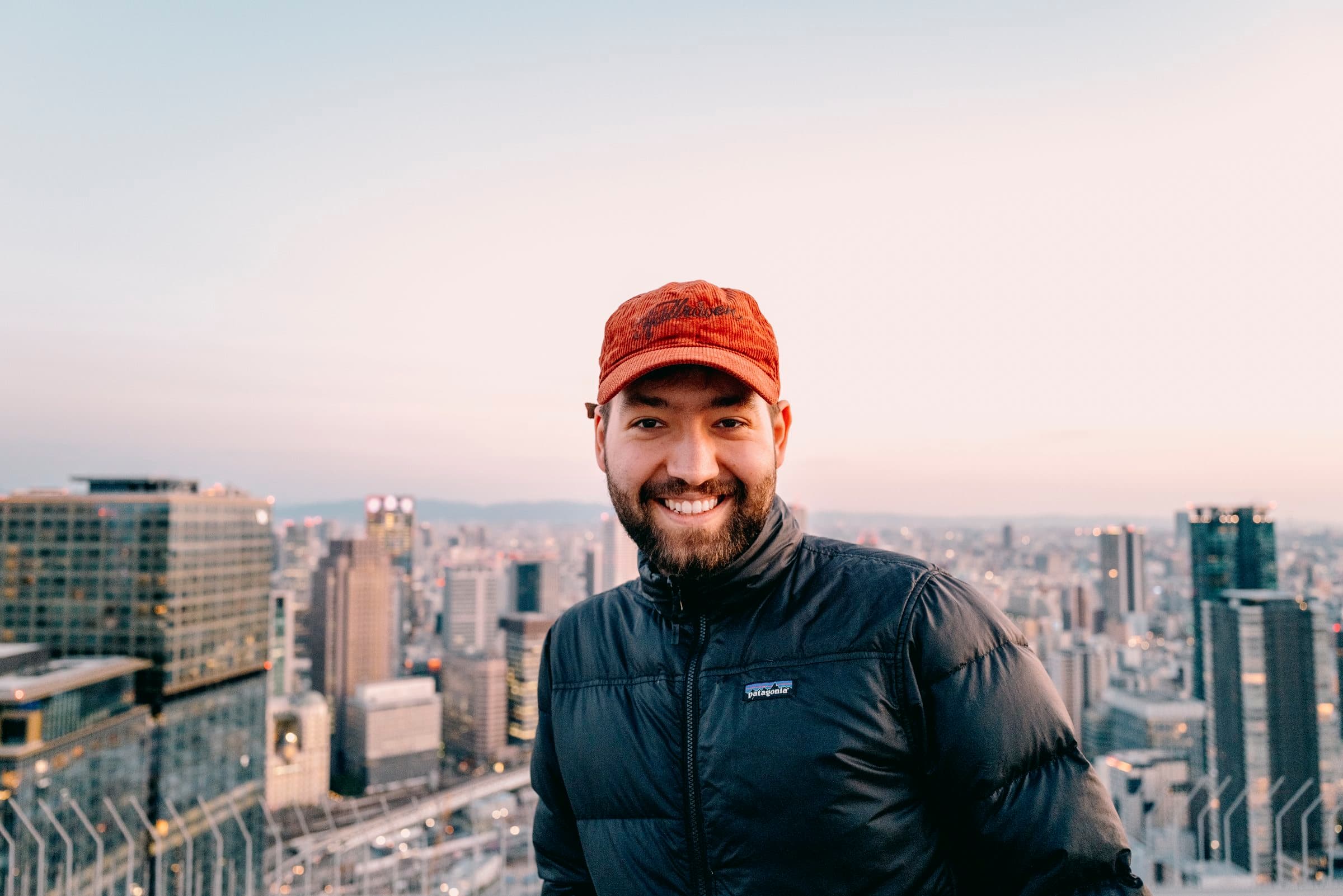 Benedict Couve in a red cap, smiling, with a city skyline at dusk behind him
