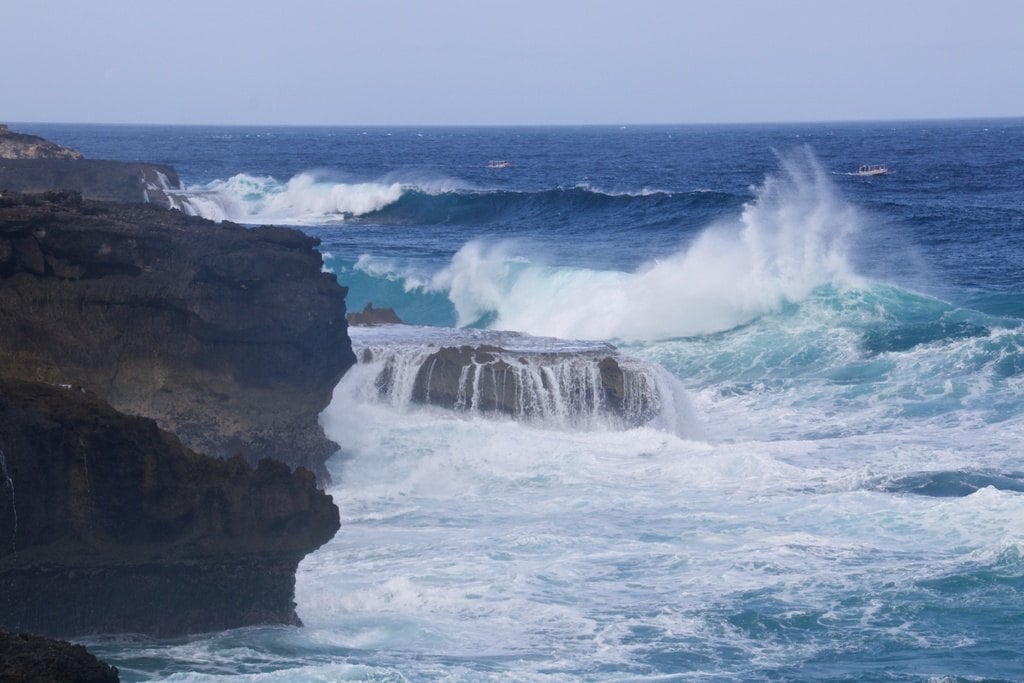Waves breaking white over black rocks under a pale sky
