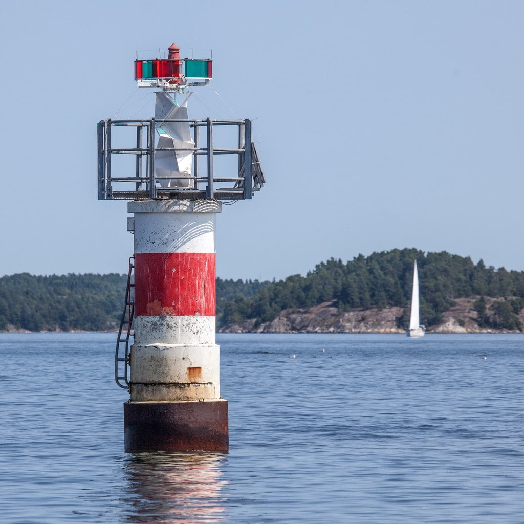 A red and white beacon tower on a rock, a sailboat behind